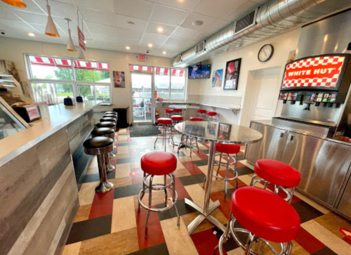 A modern diner interior with red and silver stools, a counter, and a drink station, featuring large windows and checkered decor.