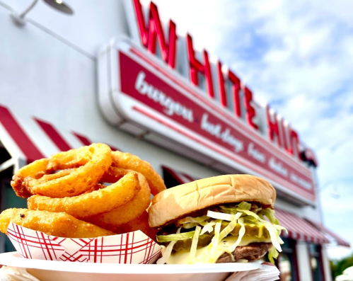 A plate with onion rings and a burger in front of a retro diner sign that reads "White Hut."