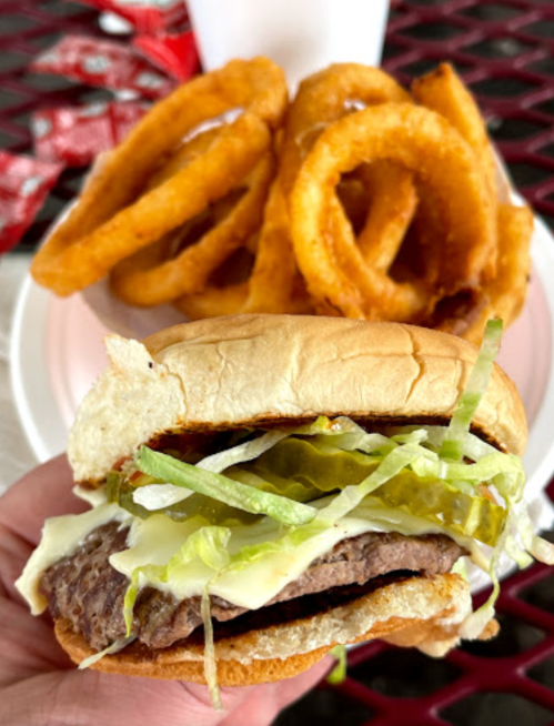 A hand holding a burger with lettuce, pickles, and mayonnaise, with a plate of onion rings in the background.
