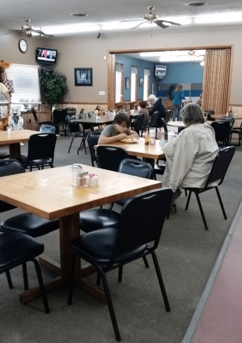 A nearly empty restaurant with wooden tables and chairs, featuring a few patrons and a TV in the background.