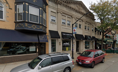Street view of a historic building with decorative architecture, flanked by shops and parked cars.