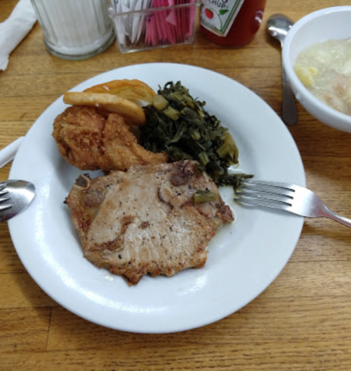 A plate of food featuring fried chicken, a pork chop, collard greens, and a bun, served with a bowl of soup.