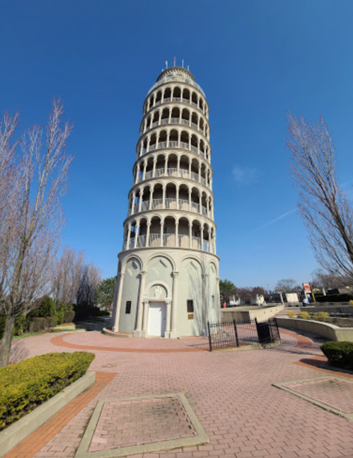 A tall, cylindrical tower resembling the Leaning Tower of Pisa, surrounded by trees and a paved walkway under a clear blue sky.