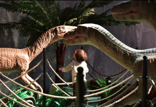 A child interacts with life-sized dinosaur models in a themed exhibit surrounded by greenery.