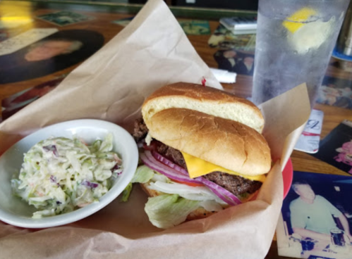 A cheeseburger with lettuce, tomato, onion, and a side of coleslaw on a wooden table with a glass of water.