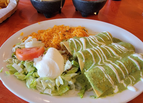A plate of green enchiladas topped with sauce, served with rice, salad, tomato, and sour cream.