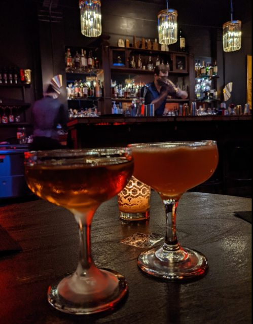 Two elegant cocktails on a bar table, with a bartender mixing drinks in a dimly lit bar in the background.
