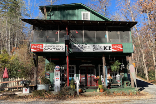 A rustic grocery store with a green exterior, featuring Coca-Cola signage and a porch surrounded by trees.
