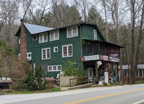 A green, two-story building with a porch, surrounded by trees, along a quiet road. Signs indicate a restaurant and shop.