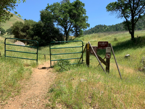 A green gate opens to a grassy trail, with a sign indicating park rules and regulations nearby.