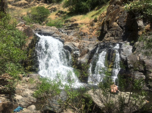 A serene waterfall cascades over rocky terrain, surrounded by lush greenery and sunlit hills.