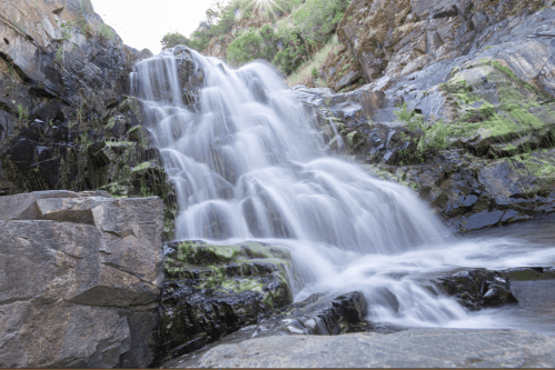 A cascading waterfall flows over dark rocks, surrounded by lush greenery and a serene natural landscape.