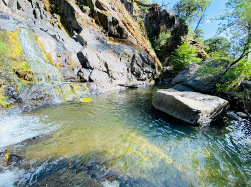 A serene natural pool surrounded by rocky cliffs and lush greenery, with a small waterfall cascading into the water.