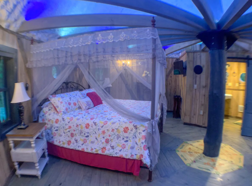 Cozy bedroom with a floral bedspread, canopy, wooden walls, and soft lighting, featuring a lamp and a bathroom door.
