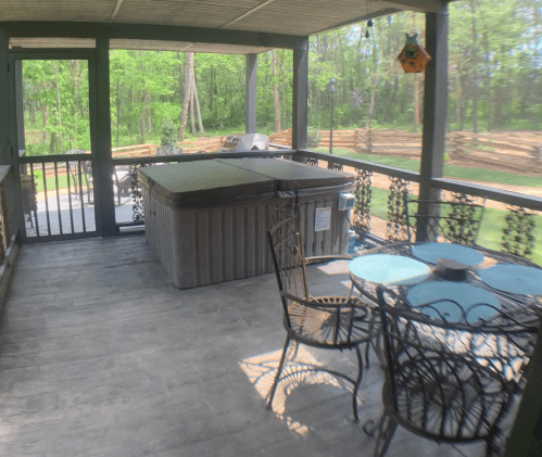A screened porch featuring a hot tub, a small table, and chairs, surrounded by greenery and wooden fencing.