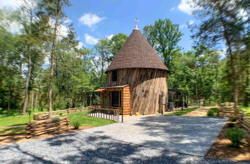 A unique, cone-shaped wooden cabin surrounded by trees and a gravel pathway. Bright blue sky above.