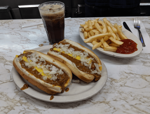 Two chili dogs topped with onions and mustard, served with a side of fries and a glass of soda on a marble table.