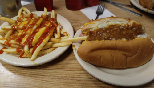 A plate with a hot dog topped with chili and a side of fries drizzled with ketchup and cheese sauce.