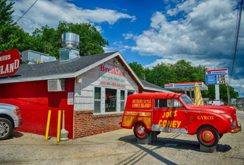 A red vintage car parked outside a diner with a sign for Joe's Coney, under a blue sky with fluffy clouds.