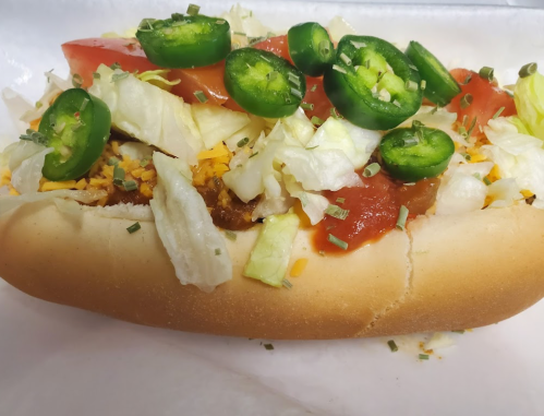 A loaded hot dog topped with lettuce, tomatoes, jalapeños, cheese, and salsa on a soft bun.