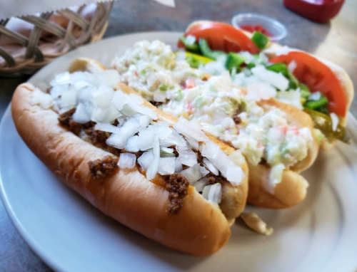 Three hot dogs on a plate, one topped with chopped onions, another with coleslaw, and the last with lettuce and tomato.