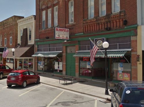 A street view of a brick building with a restaurant sign, featuring awnings and American flags, in a small town.