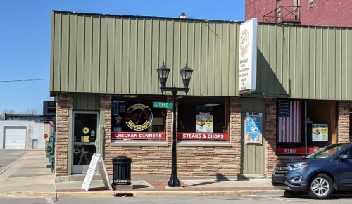 A restaurant with a green roof and stone facade, featuring signs for chicken dinners and steaks, on a sunny day.