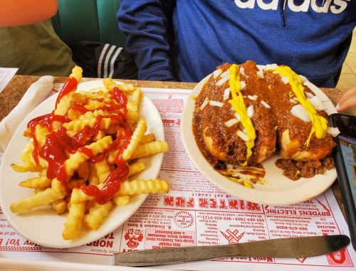 A plate of crinkle-cut fries topped with ketchup, next to a hot dog covered in chili, onions, and mustard.