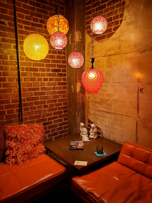 Cozy corner with orange seating, a wooden table, and colorful hanging lights against a brick wall.