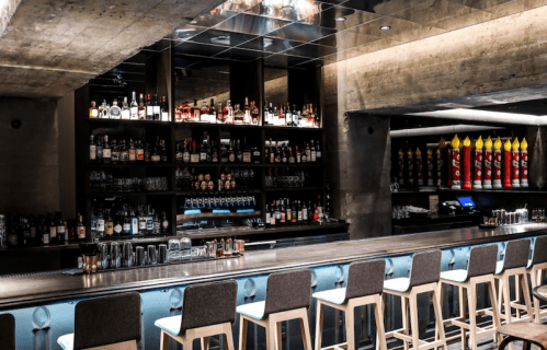 A modern bar with a sleek design, featuring a long counter and shelves stocked with various liquor bottles.