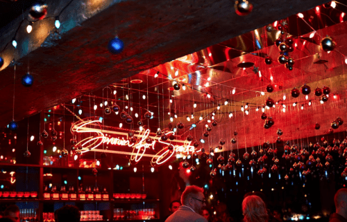 A vibrant bar scene with colorful lights, a neon sign, and decorative hanging ornaments.
