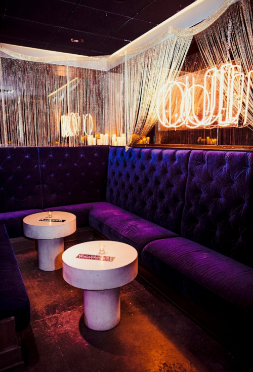 Cozy lounge area with purple velvet seating, circular tables, and soft lighting from neon fixtures and candles.