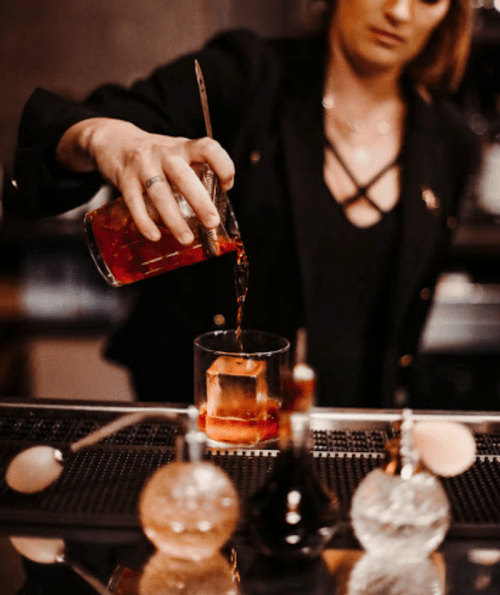 A bartender pours a cocktail into a glass, with decorative bottles in the foreground.