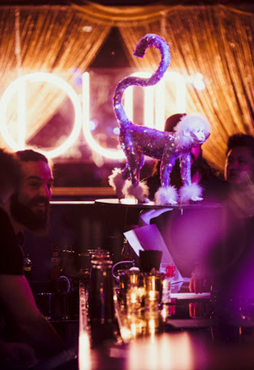 A glittery cat statue stands on a bar, with patrons enjoying drinks in a dimly lit, vibrant atmosphere.
