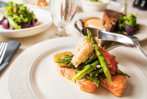 A plated dish featuring salmon topped with asparagus and greens, with side dishes and a glass of water in the background.