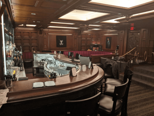A dimly lit bar with wooden paneling, a curved counter, and plush seating, featuring artwork on the walls.