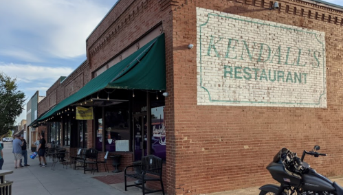 A brick building with a faded sign reading "Kendall's Restaurant," featuring outdoor seating and a motorcycle parked nearby.