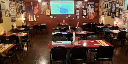 A cozy restaurant interior with red tables, black chairs, and walls adorned with framed photos and a large screen.