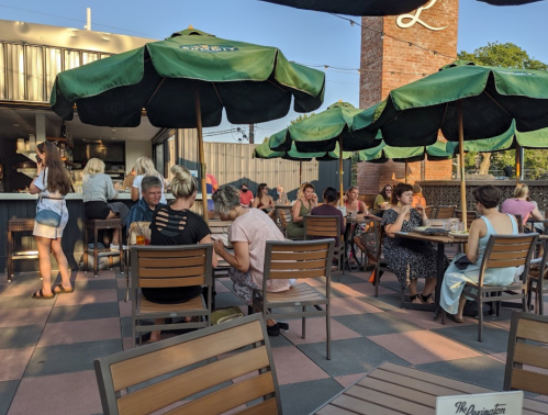 A lively outdoor dining area with people sitting under green umbrellas, enjoying food and drinks on a sunny day.