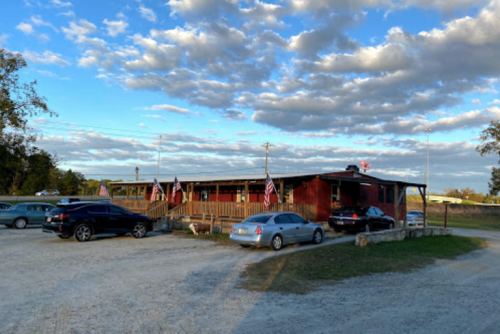 A rustic building with a wooden porch, surrounded by cars and American flags, under a blue sky with scattered clouds.