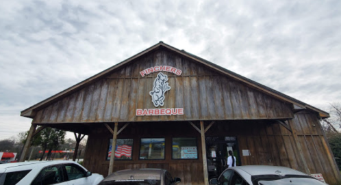Exterior of Fincher's Barbeque, a wooden building with a sign, under a cloudy sky, surrounded by parked cars.