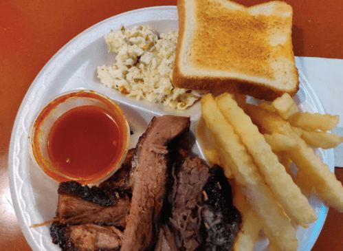 A plate with sliced brisket, crinkle-cut fries, coleslaw, toast, and a small cup of sauce.