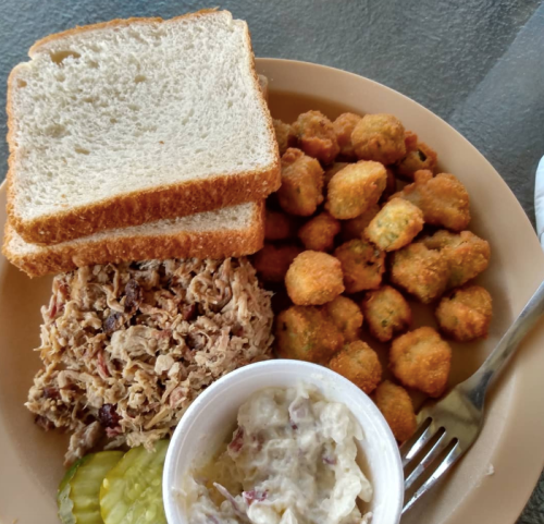 A plate with pulled pork, fried okra, coleslaw in a cup, pickles, and two slices of bread.