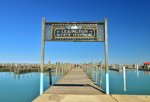Sign for Lexington State Harbor above a wooden pier extending over calm water under a clear blue sky.