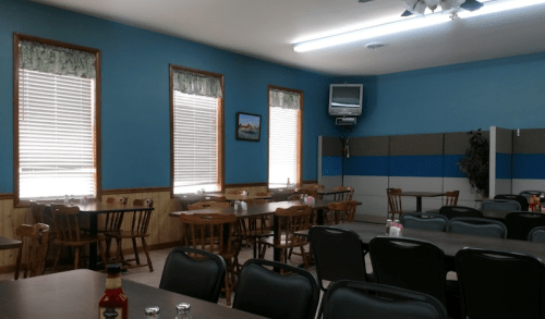 A spacious dining area with wooden tables and chairs, blue walls, and large windows with blinds.