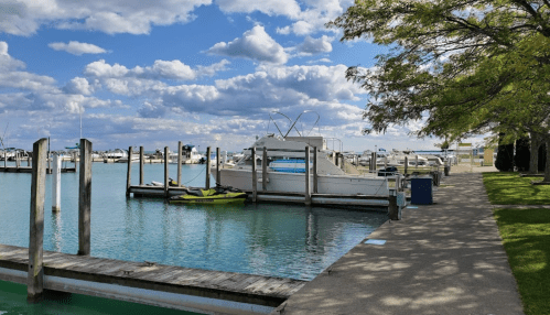 A serene marina with boats docked, surrounded by calm water and a pathway lined with trees under a partly cloudy sky.