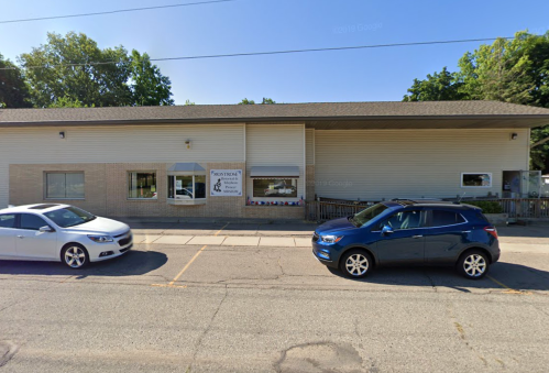 A street view of a building with large windows, featuring parked cars in front and greenery in the background.