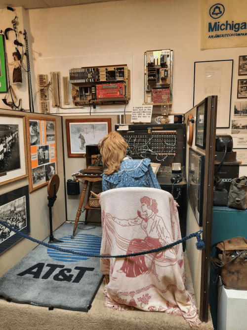 A vintage telephone operator's station with a mannequin, old equipment, and historical photos on the walls.