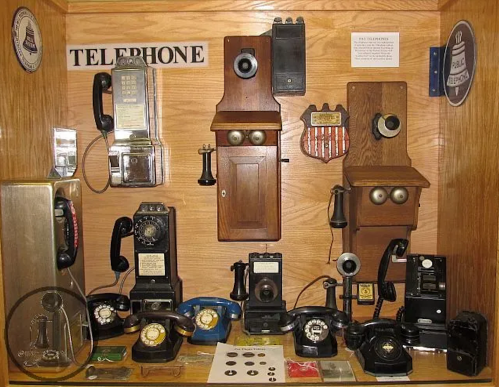 A display of various vintage telephones, including rotary and wall-mounted models, in a wooden showcase.