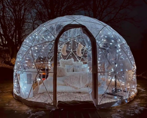 A cozy, illuminated igloo-shaped structure with soft bedding and fairy lights, set in a serene nighttime landscape.